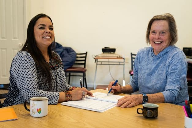 Two women smiling over a workbook at a table.