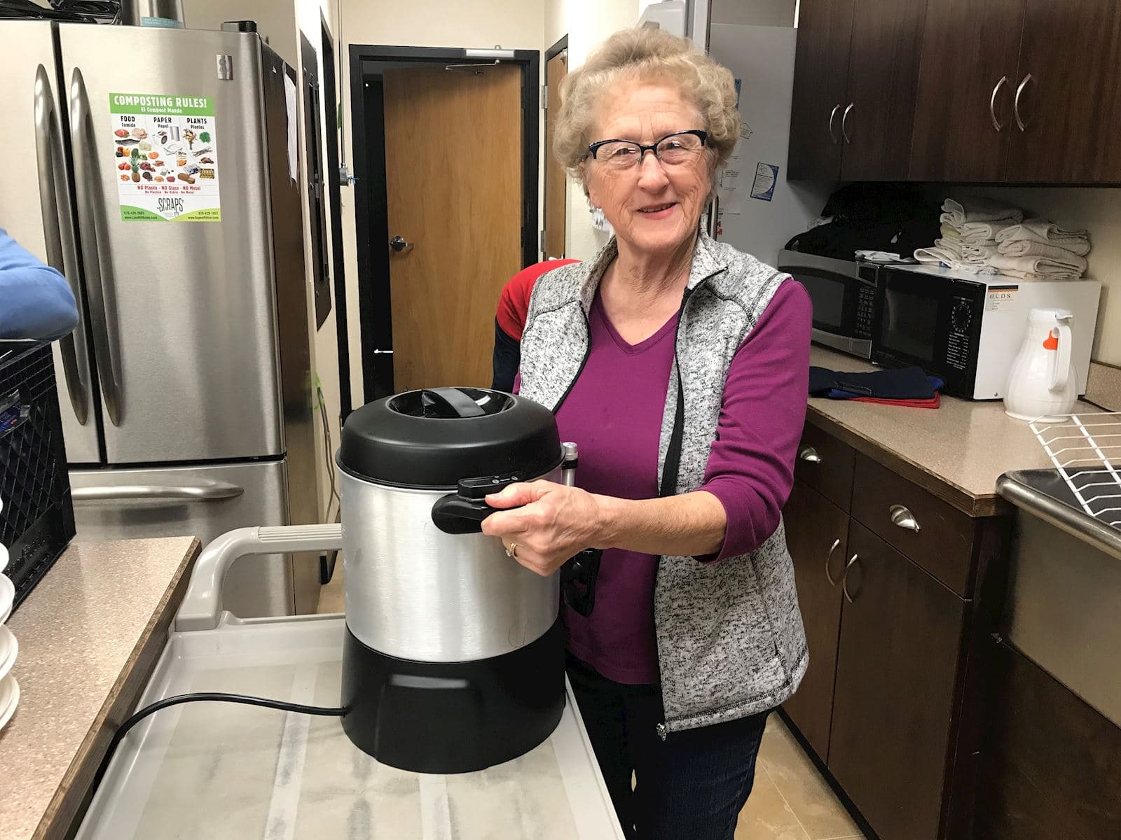 An older volunteer holding a coffee urn in a community kitchen.