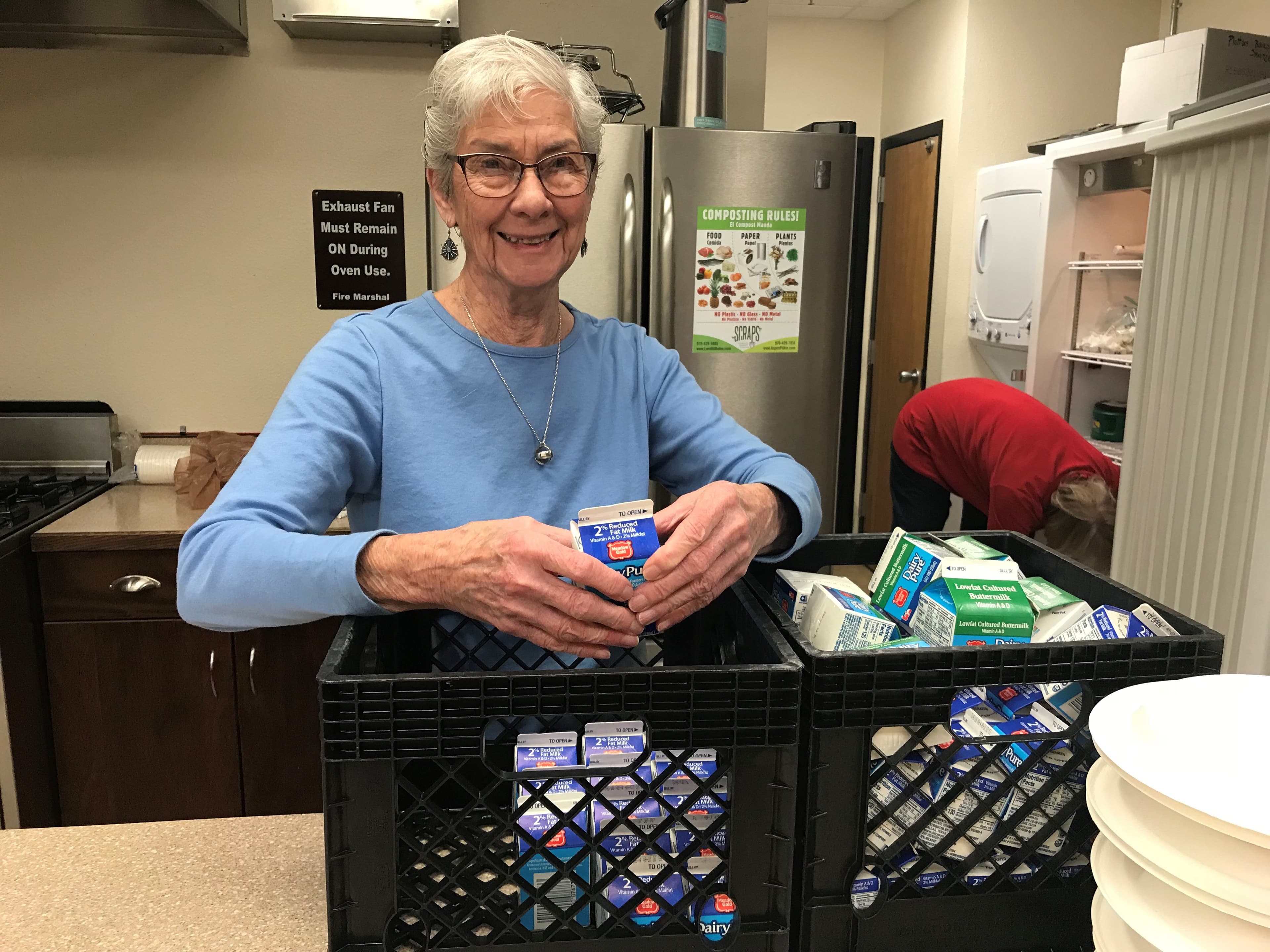A smiling volunteer packing milk cartons into crates in a community kitchen.
